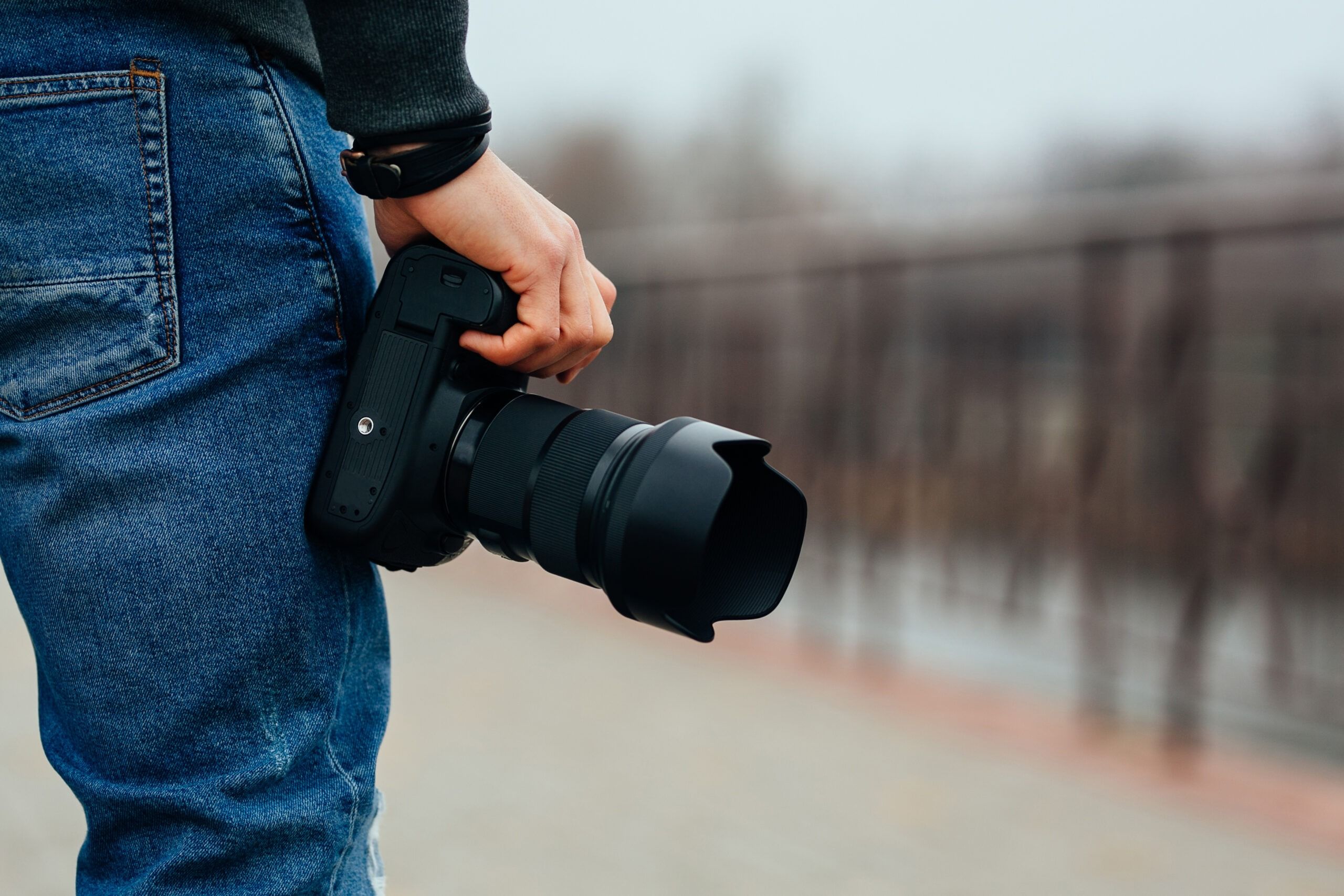 Close-up view of male hand holding professional camera on the street. Dressed in jeans.